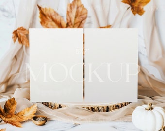 May include: A mockup featuring two 5x7 inch blank cards, ideal for showcasing designs. The cards rest on a wooden base, adorned with autumn leaves, a small white pumpkin, and a bowl of seeds. A sheer fabric drapes in the background, creating a seasonal, elegant aesthetic.