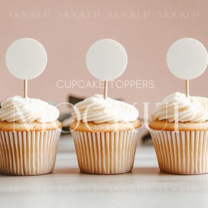 Könnte beinhalten: Drei weiße, mit Zuckerguss überzogene Cupcakes mit weißen runden Cupcake-Toppern, auf denen "CUPCAKE TOPPERS" steht, auf einem rosa Hintergrund.