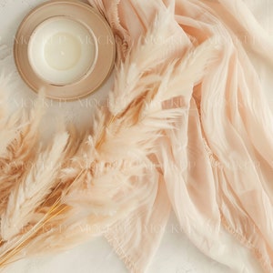 May include: A white candle in a beige ceramic dish with a beige fabric and dried pampas grass on a white background.