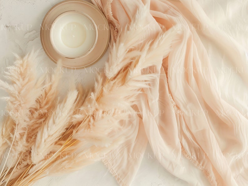 May include: A white candle in a beige ceramic dish with a beige fabric and dried pampas grass on a white background.