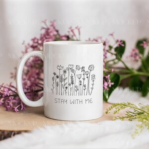 May include: White ceramic mug with a black floral line art design and the text "STAY WITH ME." The mug has a curved handle and is surrounded by purple flowers and greenery.