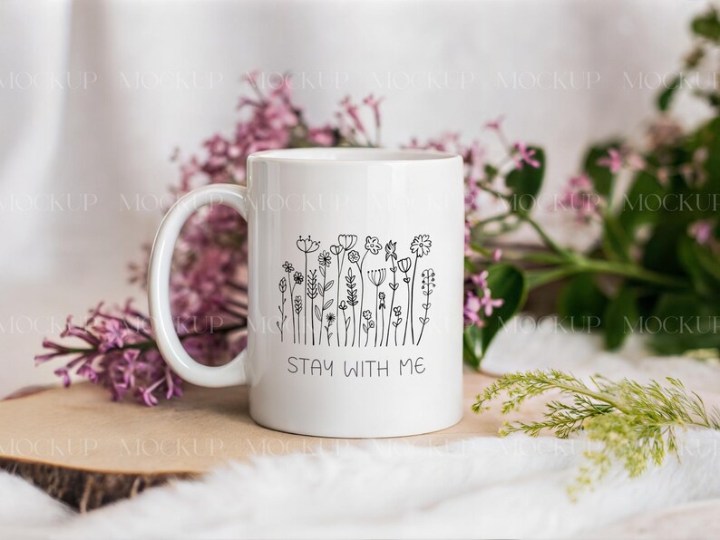 May include: White ceramic mug with a black floral line art design and the text "STAY WITH ME." The mug has a curved handle and is surrounded by purple flowers and greenery.