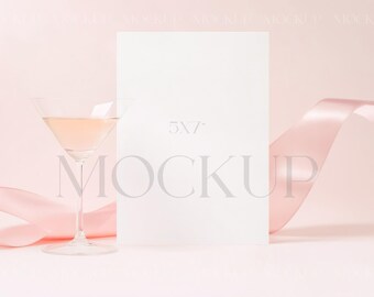 May include: A white card mockup with the text "5x7" and "MOCKUP" on a pink background with a pink ribbon and a glass of pink champagne.