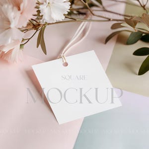 Pode incluir: Uma etiqueta quadrada branca com as palavras "SQUARE" e "MOCKUP" impressas. A etiqueta está presa a um fio branco e é colocada em um fundo pastel com flores.