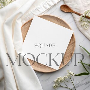 May include: A square white card with the word "SQUARE" printed on it, resting on a wooden plate. The word "MOCKUP" is printed below. The scene includes a white cloth, wooden spoon, and sprigs of white flowers and green leaves.