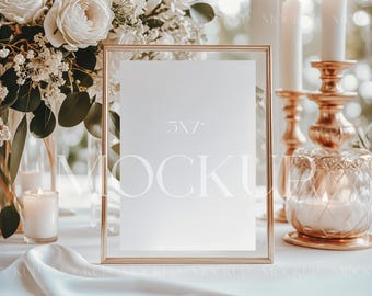 May include: A gold-framed 5x7 inch mockup with a blank white paper insert, set on a white tablecloth. The scene includes white flowers, candles, and gold candle holders, creating an elegant, romantic ambiance.