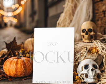Op de afbeelding: Een 5x7 inch witte kaart mockup staat te midden van een Halloween-thema arrangement. Een oranje pompoen, herfstbladeren en twee schedeldecoraties zijn rond de kaart gerangschikt. De achtergrond toont een vage straatscène met warme verlichting.