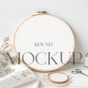 May include: A round embroidery hoop mockup with the word "ROUND" above the word "MOCKUP". The hoop is on a white surface with a white cloth, dried flowers, small scissors, and jars with contents.