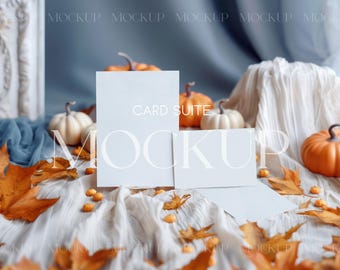 May include: A vertical rectangular card and a smaller horizontal rectangular card displayed on a white fabric surface. The cards are surrounded by orange pumpkins, white pumpkins, and scattered dried autumn leaves in shades of orange and brown.