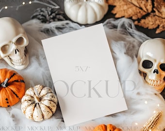 Op de afbeelding: Een Halloween-thema mockup met een blanco kaart van 13 x 18 cm, omringd door pompoenen, schedels en herfstbladeren. De pompoenen zijn oranje en wit, en de schedels zijn wit. De scène speelt zich af op een grijze ondergrond met witte stof.