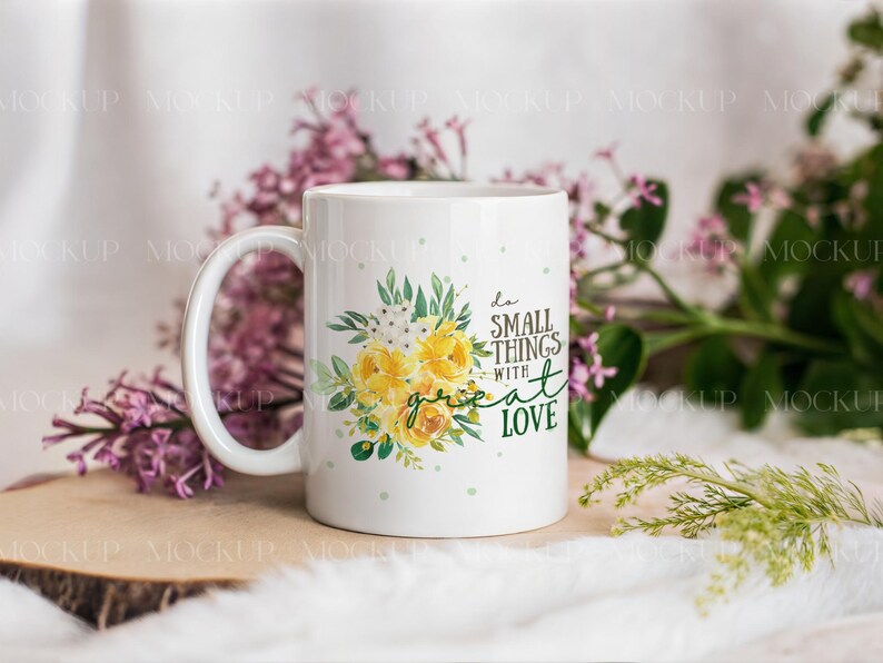 May include: White ceramic mug with a floral design featuring yellow roses and green leaves. The mug has the text "do small things with great love" in a mix of fonts. The mug is surrounded by purple flowers and greenery.