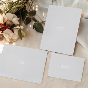 May include: A mockup featuring three white rectangular cards of varying sizes: 5x7 inches, 7x5 inches, and 5x3.5 inches. The cards are arranged on a light-colored fabric, with a floral arrangement of white and red flowers and green leaves in the background.