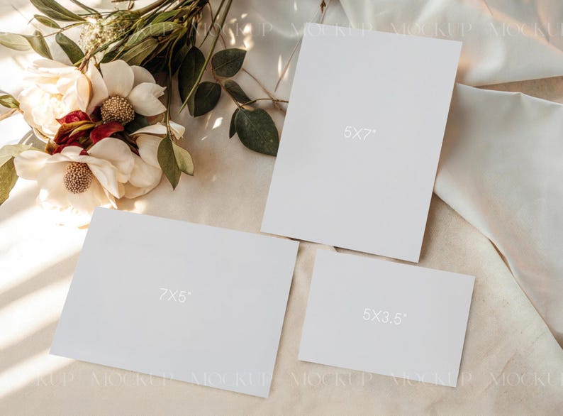 May include: A mockup featuring three white rectangular cards of varying sizes: 5x7 inches, 7x5 inches, and 5x3.5 inches. The cards are arranged on a light-colored fabric, with a floral arrangement of white and red flowers and green leaves in the background.