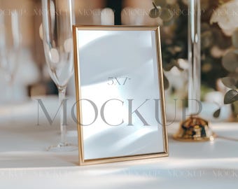 May include: A gold-colored picture frame with a white mat and a clear glass front. The frame is sitting on a white tablecloth and has the text "5x7" and "MOCKUP" printed on it.