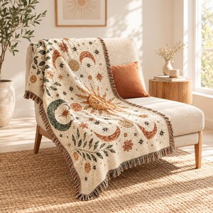 Peut inclure: Un jeté de lit tissé de couleur crème orné d'un motif répétitif de soleils, de croissants de lune, d'étoiles et de branches feuillues dans des tons terre cuite, vert foncé et or. La couverture présente des bords à franges sur un fauteuil.
