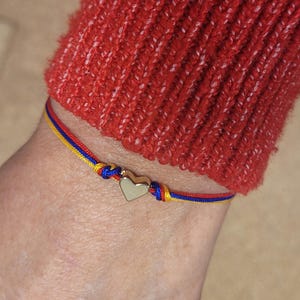 May include: A gold heart charm bracelet with red, yellow, and blue cord. The bracelet is knotted and worn on a wrist, with a red sweater in the background.