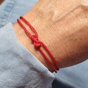 May include: A red string bracelet tied with a knot. The bracelet is worn on a wrist, with the red string contrasting against the skin tone and a blue denim shirt sleeve in the background. The knot is centered, and the string is thin.