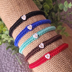 May include: Five colorful friendship bracelets with heart-shaped charms. Each charm has a different letter, spelling out the word 'R.O.J.E.K'.