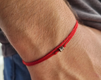 May include: Red string bracelet with a small, square, silver-colored bead. The bracelet is worn on a person's wrist. The background is blurred, showing a gray shirt and blue jeans.