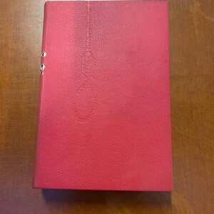 May include: A red binder with a textured surface. The binder has three metal rings on the spine and a subtle embossed design on the front cover. The binder is rectangular and appears to be closed.