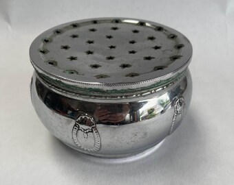 May include: A silver-colored, round container with a perforated lid. The lid features star-shaped cutouts. The container has a bulbous body with decorative oval designs. The metal has a slightly tarnished appearance.