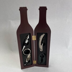 Pode incluir: Uma caixa de madeira em forma de garrafa de vinho com tampa articulada, contendo um saca-rolhas, uma rolha de garrafa e um despejador. A caixa é de cor castanho escuro e tem um interior preto.