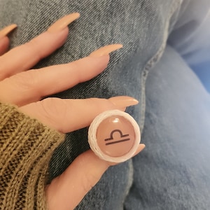 Op de afbeelding: Een ronde ring met het sterrenbeeld Weegschaal, met een witte geweven rand en een roze achtergrond. Het Weegschaal-symbool is zwart. De ring wordt getoond tegen een achtergrond van blauwe spijkerstof en een groene trui.