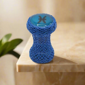 May include: A blue woven glass holder with a rounded top. The top features a blue background with a black Pisces symbol. The holder is made of woven blue material and sits on a light-colored surface.
