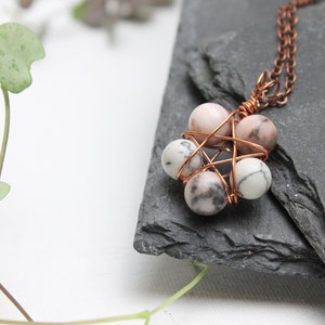 Op de afbeelding: Een ketting met een hanger van koperdraad gewikkeld rond vijf roze en witte gemarmerde stenen. De ketting ligt op een donkergrijs leisteen oppervlak.