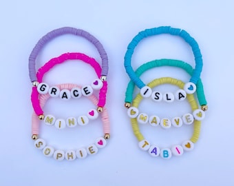 May include: Set of six colorful beaded bracelets with names spelled out in white beads with gold lettering. The names are "Grace", "Mini", "Sophie", "Isla", "Maeve", and "Tabi".