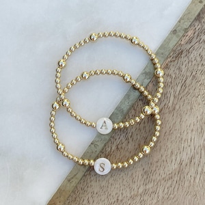 May include: Two gold beaded bracelets with white round charms. The charms have the letters "A" and "S" engraved on them.