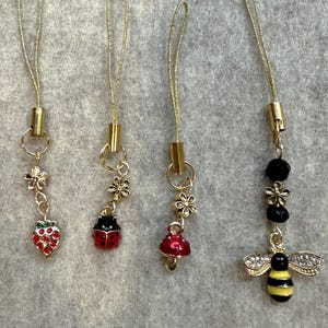 May include: Four decorative charms with gold-colored cords and attachments. The charms feature a red strawberry, a ladybug, a red mushroom, and a bee with black and yellow stripes. Each charm has a gold-colored flower accent.