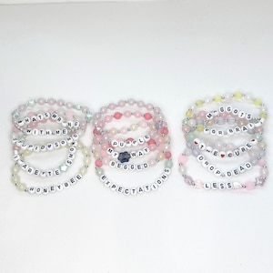 May include: Three clusters of beaded bracelets featuring white letter beads spelling phrases like WHATS WRONG WITH ME, STUPID SONG, SMOKING, HONEYBEE, PURPLE, MY WAY, BEGGED, EXPECTATIONS, MAGGOTS FOR BRAINS, THE CURE, and DROP DEAD LESS.