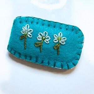 May include: A rectangular turquoise felt hair clip featuring three embroidered white flowers with green stems and small green bead centers. The edges of the felt are finished with a decorative blanket stitch in matching turquoise thread.