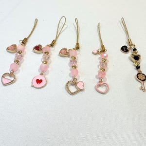 May include: Five phone charms featuring gold-tone hardware, pink and clear faceted beads, and small gold heart spacers. Four charms include pink enamel heart-shaped pendants, while the fifth charm features a black heart and a gold key pendant.