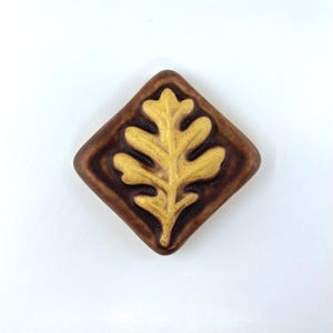 May include: A diamond-shaped ceramic tile featuring a raised, embossed oak leaf design in a mustard yellow glaze. The background of the tile is a deep, dark brown with a smooth, glossy finish that contrasts with the textured yellow leaf center.