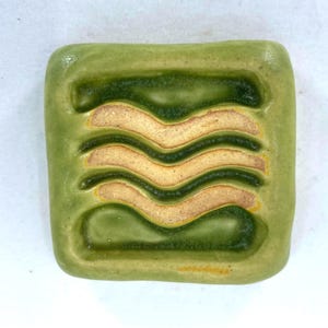 May include: A green ceramic tile with a wavy pattern in a lighter shade of yellow. The tile has a slightly raised edge.