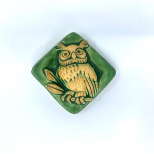 May include: A diamond-shaped ceramic tile featuring a raised, light tan owl relief centered on a glossy, deep green background. The owl is depicted with detailed feathers and large eyes, perched on a branch with two visible leaves.