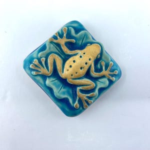 May include: A square ceramic tile with rounded corners featuring a raised, golden-tan frog relief centered on a vibrant, glossy teal blue background. The frog is depicted in a sprawling position with detailed toes and a textured, spotted back.