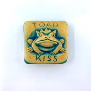 May include: A square ceramic tile featuring a raised relief of a toad with a crown. The toad is colored in a light tan, while the background and edges are a deep teal blue. The text TOAD KISS is embossed in the tile above and below the toad.