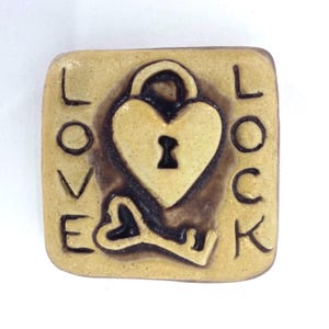 Puede incluir: Azulejo cuadrado de cerámica beige con un diseño de cerradura y llave en forma de corazón, con las palabras "LOVE" y "LOCK" a cada lado. La cerradura y la llave son de color marrón oscuro, y el azulejo tiene una superficie ligeramente texturizada.