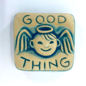 May include: A square ceramic tile with a smiling angel face, halo, and wings, in a light tan color with teal blue accents. The words "GOOD THING" are inscribed above and below the angel illustration.