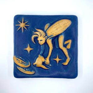 May include: A square blue ceramic tile featuring a raised relief design of a golden, humanoid figure with flowing hair bending over to touch a golden object. The scene includes three golden four-pointed stars and two golden oval shapes.