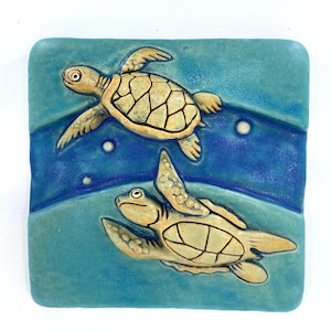 May include: A square ceramic tile featuring two swimming sea turtles. The turtles have tan shells with a grid pattern and are set against a blue background with wavy lines and bubbles. The tile has a turquoise border.