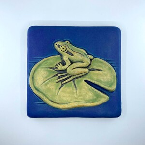 May include: A square ceramic tile featuring a relief design of a light green frog sitting on a pale green lily pad. The background is a deep, solid blue, and the entire piece has a matte finish with visible dark outlines defining the frog and leaf.