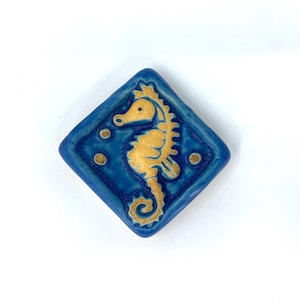 May include: A diamond-shaped ceramic tile featuring a raised, golden-yellow seahorse relief centered on a deep blue background. The design includes three small, circular golden-yellow bubbles floating near the seahorse, all set against a smooth, glossy finish.