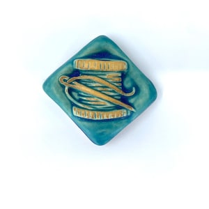 May include: A diamond-shaped ceramic button in a glossy teal glaze featuring a raised, tan-colored relief design of a spool of thread with a needle resting across it. The surface has a smooth, reflective finish with subtle color variations.