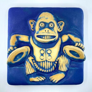 Puede incluir: Azulejo de cerámica azul con diseño de mono. El mono está sentado, con un chaleco amarillo y sosteniendo platillos amarillos. El azulejo es cuadrado y los detalles del mono son visibles.