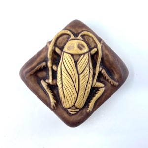 May include: A brown and tan ceramic tile with a detailed relief of a cockroach. The insect is centered on a diamond-shaped tile, with the cockroach in tan and the tile in a darker brown.