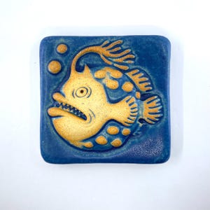 May include: A square ceramic tile featuring a raised, textured golden anglerfish design against a deep blue background. The fish has a prominent lure, sharp teeth, and rounded spots, accompanied by two small golden bubbles in the upper left corner.